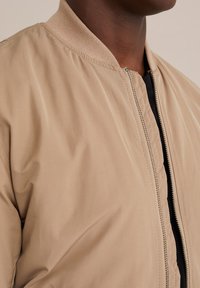 Světle béžová bomber bunda s žebrovaným límcem, předním zipem a hladkým, mírně lesklým materiálem. Vyznačuje se jednoduchým, čistým designem.