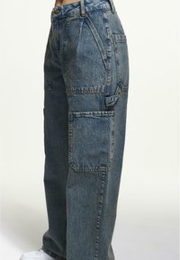 Jeans cargo en denim azul desgastado, con pierna recta, múltiples bolsillos laterales y costuras reforzadas para mayor durabilidad. Zapatilla blanca visible abajo.