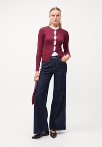Cardigan lung cu mâneci lungi de culoare burgund, cu detalii cu nasturi, combinat cu blugi evazați de culoare albastru închis. Pantofi negri cu accente decorative și o geantă roșie.