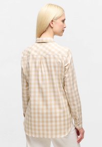 Woman with long blonde hair wearing a beige and white checkered long-sleeve shirt, facing away against a plain white background.