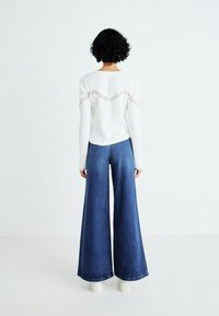 Witte geribbelde langemouwen top met kantdetails en blauwe denim wijde broek. Model draagt witte plateau schoenen en staat tegen een effen achtergrond.