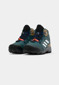 Botas de caminhada verdes de cano médio com solas de borracha pretas, riscas brancas e atacadores azuis. Apresentam realces castanhos e um material superior texturizado.
