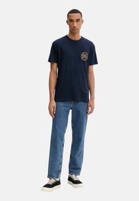 Camiseta de algodón azul marino con cuello redondo y estampado gráfico dorado en el lado izquierdo del pecho, combinada con jeans de mezclilla azul claro y zapatillas negras.