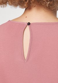 Kortärmad, rosa blus med en nyckelhålsöppning bak som stängs med en svart knapp. Strukturerad tyg, rund halsringning och sydda kanter.