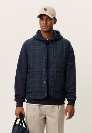 Veste matelassée avec motif à carreaux bleu marine et vert, fermetures à pression et poches latérales, portée sur un sweat à capuche bleu marine avec cordon de serrage.