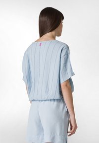 Top corto de manga corta en azul claro con rayas verticales y un escote amplio, con una pequeña etiqueta rosa en la parte posterior. Combinado con pantalones cortos azul claro.