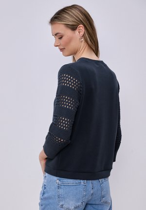 Frau trägt einen schwarzen Pullover mit strukturierten, offen gestrickten Ärmeln und hellblauen Jeans, steht seitlich vor einem einfarbigen Hintergrund.