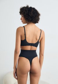 Black lace-back bralette with adjustable straps, paired with high-waisted black thong. Smooth fabric, scalloped detailing on the bra edges.