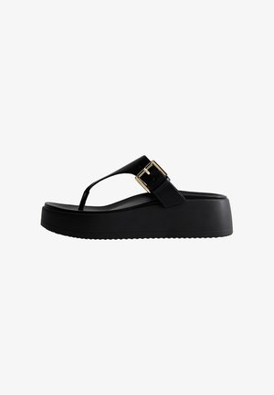 Sandalia tipo flip-flop negra con suela gruesa, tira tipo tanga y hebilla grande dorada en el lateral.