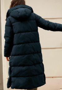 Manteau rembourré bleu marine avec capuche, présentant des coutures horizontales, des manches longues et des fentes latérales près de l'ourlet pour plus de facilité de mouvement.