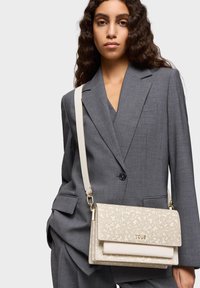 Beige structured handbag with a textured floral pattern, featuring a cream strap and gold "TOUS" logo, complemented by a gray suit.