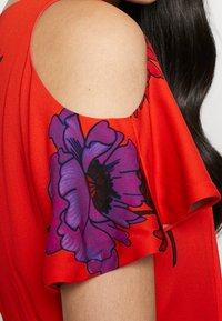 Femme portant un haut rouge à épaules dénudées avec un grand imprimé floral violet sur la manche, cheveux longs et foncés visibles sur un fond blanc.