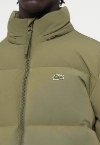 Zeleni puffer jakna s visokim ovratnikom i prednjim centar zatvaračem. Sadrži izvezeni logotip na lijevoj dojci i glatku teksturu tkanine.