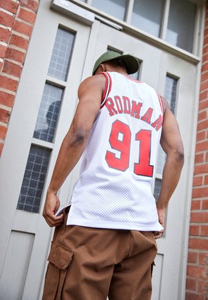 Maillot de basket-ball en maille blanche avec des accents rouges, portant "RODMAN" et "91" au dos. Short cargo marron, porte avec des fenêtres à panneaux en arrière-plan.