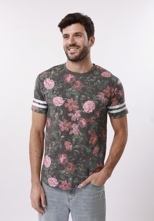 Lächelnder Mann mit dunklem Haar, der ein graues T-Shirt mit Blumenmuster und weißen Streifen an den Ärmeln sowie hellblaue Jeans trägt, eine Hand in der Hosentasche.
