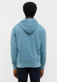Personne avec des cheveux bouclés courts portant un sweat à capuche bleu clair et un jean noir, de dos devant un fond blanc uni.