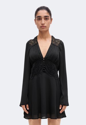 Un mannequin se tient devant un fond clair uni, vêtu d’une robe noire à manches longues avec des empiècements en dentelle, un décolleté en V profond et des boutons sur le devant.