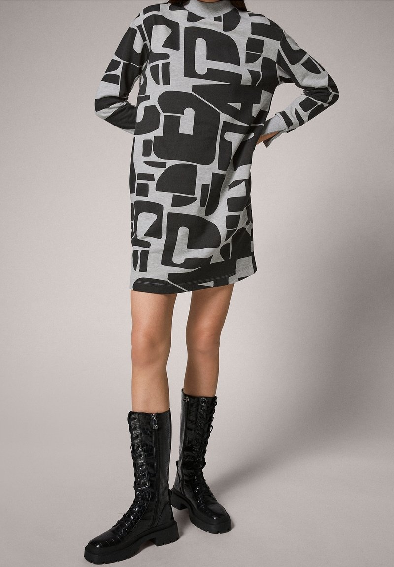 Robe grise avec de grandes formes abstraites noires, manches longues et encolure ronde. Associée à des bottes noires montantes à lacets jusqu'aux genoux.