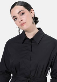 Donna con capelli corti scuri che indossa orecchini in argento a catena e un vestito nero con colletto e vita legata, su sfondo bianco uniforme.