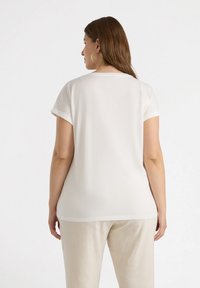Top blanco de manga corta con cuello redondo y puños enrollados, hecho de una tela suave. Presentado en un modelo que lleva pantalones de color claro.