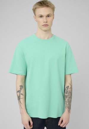 Jeune homme aux cheveux blonds courts portant un t-shirt vert menthe, montrant des tatouages floraux et papillons sur les deux avant-bras.