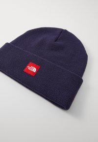 Berretto in maglia blu navy con orlo arrotolato, caratterizzato da una toppa quadrata rossa con testo e logo bianchi. Texture morbida e design semplice.