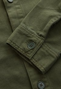 Gros plan du poignet d'une chemise à manches longues vert olive avec une lanière cousue à bouton et des boutons assortis sur tissu texturé.