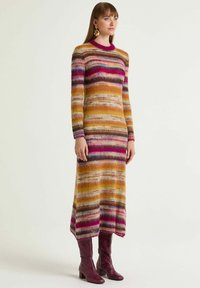 Strickkleid mit mehrfarbigen Streifen in Gelb, Rosa und Lila, mit Rundhalsausschnitt und langen Ärmeln. Kombiniert mit burgunderroten, strukturierten Stiefeln.