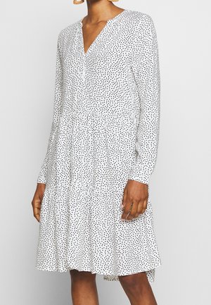 Woman wearing a white long-sleeve dress with black polka dots and a tiered skirt, standing against a plain light background.