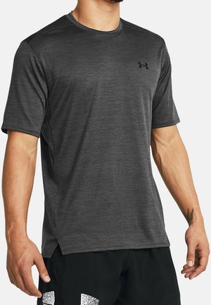 Mężczyzna ubrany w ciemnoszarą sportową koszulkę z krótkim rękawem z logo Under Armour na piersi oraz czarne szorty z białym wzorem.