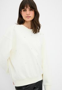 Krémová mikina s volným střihem, zdobená rozptýlenými diamantovými aplikacemi a žebrovanými manžetami a lemem. Hladká textilie.