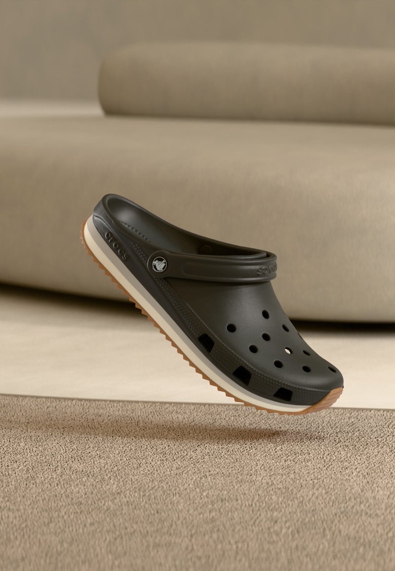 Sabot Croslite noir avec des trous de ventilation et une semelle en caoutchouc couleur naturelle, flottant au-dessus d’un tapis beige et d’un canapé en arrière-plan.
