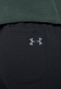 Detailní záběr na černou látkovou kapsu kalhot s vybledlým logem Under Armour, pod texturovaným tmavě zeleným oděvem.