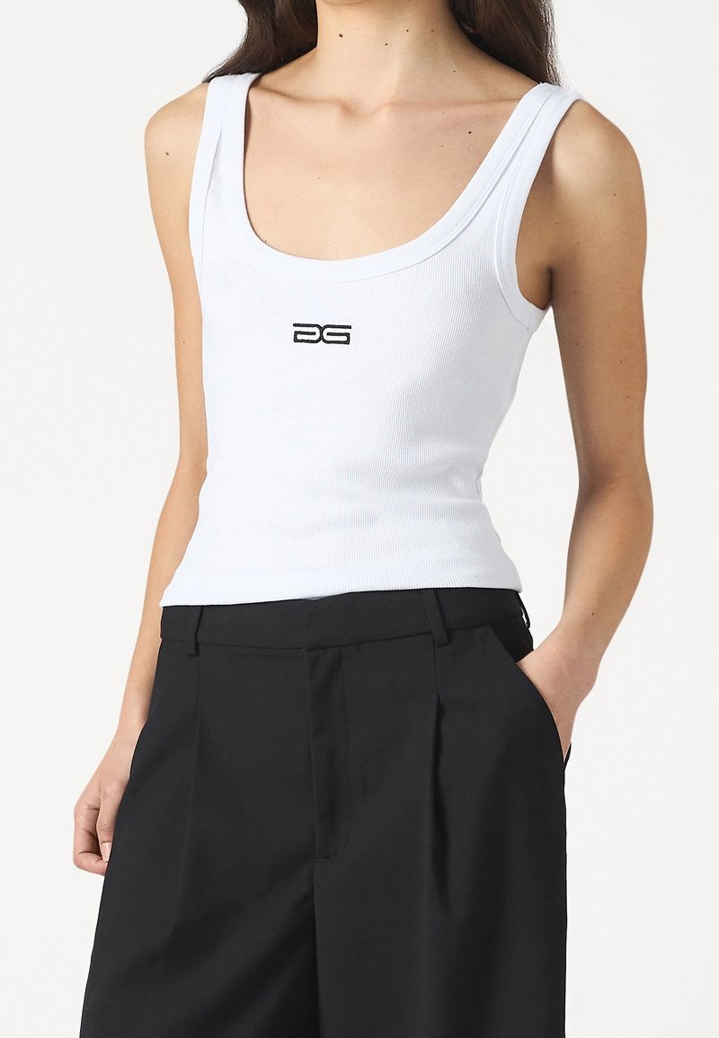 Kvinna klädd i en vit ärmlös ribbad topp med en liten svart logotyp på bröstet och högmidjade svarta byxor med händerna i fickorna.