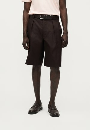 Άνδρας που φοράει μαύρα ραμμένα στα μέτρα του βερμούδα μέχρι το γόνατο με πιέτες, μαύρη ζώνη, καφέ επίσημα παπούτσια και ροζ ανοιχτό κοντομάνικο πουκάμισο.