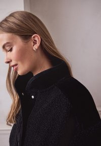 Veste en polaire noire avec un col montant, caractérisée par une texture douce, des boutons-pression argentés et des accents de tissu contrastants sur les épaules.