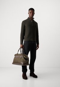 Sudadera de cuello con cremallera en verde oliva, jeans oscuros y botas de ante marrón. Sosteniendo un gran bolso verde oliva con detalles marrones y un bolsillo frontal.