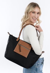 Borsa tote nera con una tasca frontale in pelle marrone, dettagli con zip e doppio manico. Il tessuto è liscio con una finitura opaca.
