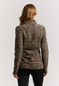 Woman with long hair wearing a leopard print long-sleeve top and black pants, facing away from the camera against a plain background.
