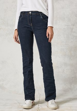 Donkerblauwe denim jeans met een rechte snit, voorzakken, zichtbare stiksels en een logo-detail bij de taille, gecombineerd met witte sneakers.