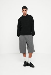 Pull en tricot noir avec un col rond, associé à un short plissé gris. Le mannequin porte des mocassins noirs et des chaussettes blanches à la cheville.