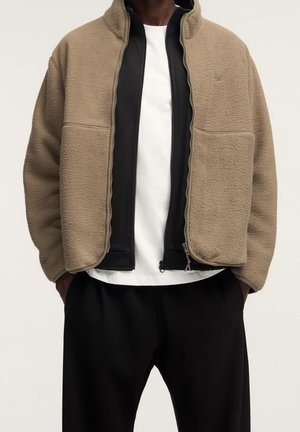 Mand iført lagdelt beige fleecejakke over sort lynlåsjakke og hvid skjorte, med sorte bukser, hænderne i lommerne, neutral baggrund.