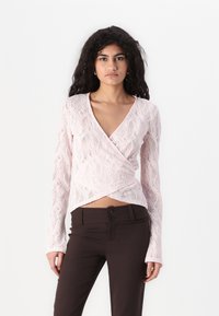 Hellrosa Spitzenoberteil mit langen Ärmeln und Wickeldesign, das einen V-Ausschnitt und ein strukturiertes Blumenmuster aufweist. Kombiniert mit braunen, eng anliegenden Hosen.