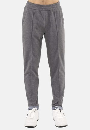 Homme portant un pantalon de survêtement gris et des baskets blanches, debout devant un fond uni, montré de la taille jusqu'aux pieds.