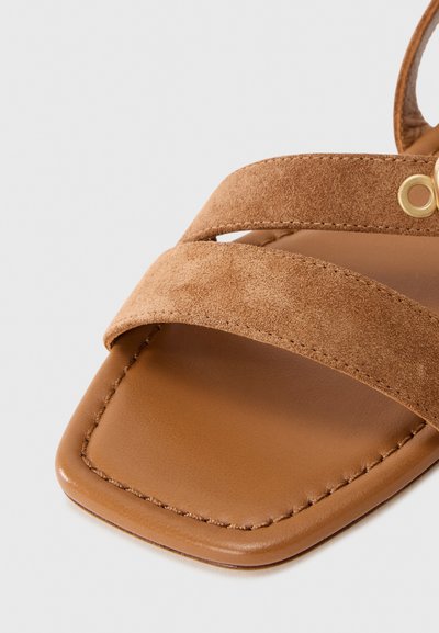 Bruna mockaskinnsandaler med platta sulor som har två korsande remmar, en slät läderfotbädd och fyrkantig tå. Guld dekorativ detalj nära remmen.