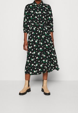 Kvinde iført en sort midi-kjole med hvidt og grønt blomsterprint, taljefremhævet, med trekvartlange ærmer og beige ankelstøvler med sorte såler.