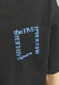 Camiseta de algodón negra con texto bordado en azul dentro de un contorno cuadrado. El texto dice "the FIVE B*ROUGHS Atelier Originals."