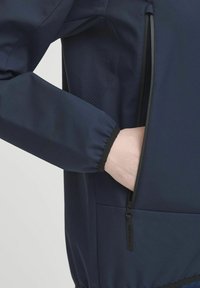 Marineblaues Jacket aus glattem Material, mit einer Reißverschlusstasche und Netzbelüftungsdetails. Die Bündchen sind gerafft; das Logo ist auf dem Ärmel sichtbar.