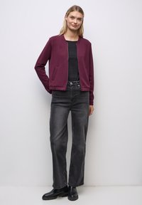 Burgundy bomberjacka med framdragkedja, ribbad nederkant och sidofickor, tillsammans med svarta jeans och ankelskor. Neutral bakgrund.