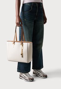 Un sac fourre-tout blanc avec des poignées en cuir marron, présentant des éléments en or, un charme en forme de pendentif et un design structuré. Porté avec un jean large bleu et des baskets.